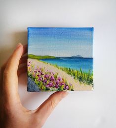 a person holding up a small painting on a white surface with water and flowers in the foreground