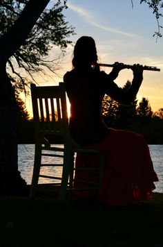 a woman is sitting on a bench playing the flute in front of a lake at sunset