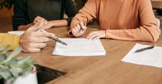 two people sitting at a table with papers and pen in their hands, one person is writing something on the paper