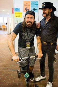 two men standing next to each other with one on top of the bike while another stands behind them