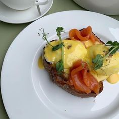 a white plate topped with an open face sandwich covered in cheese and toppings next to a cup of coffee