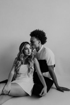 a man and woman sitting next to each other in black and white photo with one kissing the other's cheek