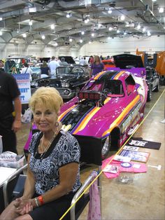 a woman sitting in front of a pink car at a car show with other cars on display