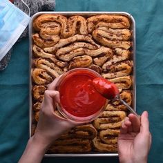 a person holding a spoon in front of a pan filled with cinnamon rolls and ketchup