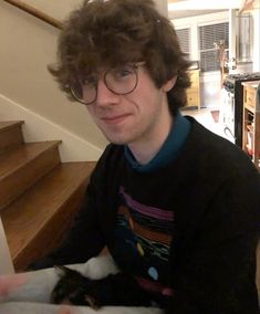 a man with glasses holding a cat in his lap while sitting on the floor next to stairs