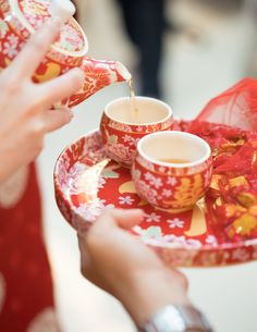 a woman pouring tea into two cups on a red and yellow plate with gold trim