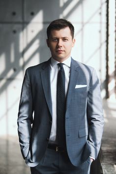 a man wearing a suit and tie standing in front of a window with his hands in his pockets