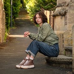 a young woman sitting on the side of a road next to a stone building and trees