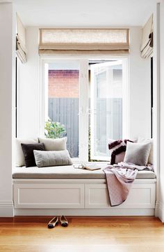 a window seat with pillows on it in front of a wooden floor and white walls