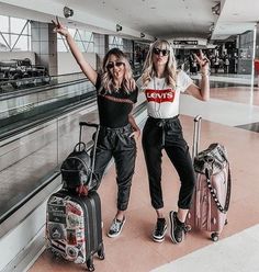 two women standing next to each other with their arms in the air and luggage behind them