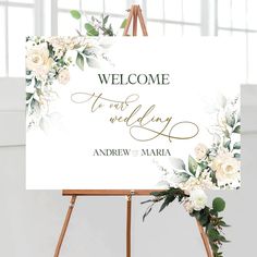 a welcome sign with white flowers and greenery on an easel in front of a window