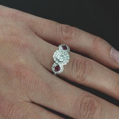 Edwardian Three Stone Moissanite Ruby Flower Halo Diamond Forever One Floral Engagement Ring Ornate and romantic three stone engagement ring made of 14K white gold. Ring features two round, bezel set red rubies (3.5mm each, .38CT) on either side of a round, 5mm (~.5ct) Forever One Moissanite center stone. All three stones are surrounded by flower shape halos of pave-set, round diamonds (.36cttw, G-VS quality). High polish, smooth sides and edges, cut-out baskets support bigger stones. Dimensions White Gold Three Stone Proposal Rings, White Gold Three Stone Ring For Proposal, Fine Jewelry Three Stone Rings For Proposal, Anniversary Ruby Ring With Center Stone In Round Band, Moissanite Cluster Ring With Rose Cut Diamonds For Anniversary, Ruby Diamond Ring With Rose Cut For Proposal, White Gold Three Stone Diamond Ring For Proposal, Wedding Cluster Diamond Ring With Gemstone, Ruby Anniversary Ring With Center Stone