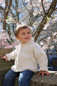 a little boy sitting on top of a stone wall next to trees with pink flowers