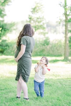 a pregnant woman standing next to a little boy in the grass with trees behind her