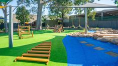 An outdoor childcare sheltered by grey sail shades and is divided half of the floor by green and the other by blue grass. A stair case looking wooden platform lies on the grass and there is a wooden climbing wall in the back. Climbing Equipment, Water Play