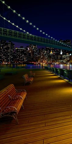 two benches sitting on top of a wooden deck next to the water at night with city lights in the background