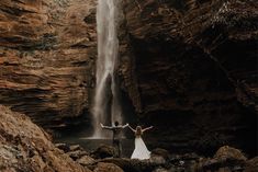 a bride and groom standing in front of a waterfall