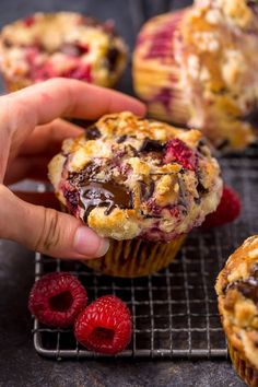 a hand holding a muffin with chocolate and raspberry toppings next to some raspberries