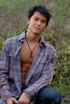 a shirtless young man sitting on the ground in front of some grass and bushes