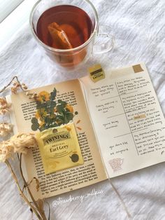 an open book next to a cup of tea and dried flowers on a white table cloth