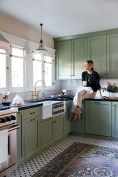 a woman sitting on top of a kitchen counter next to an oven and sink with green cabinets