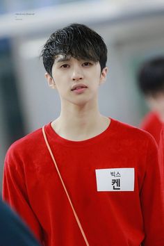 a young man with black hair wearing a red shirt and looking at the camera while standing in front of other people