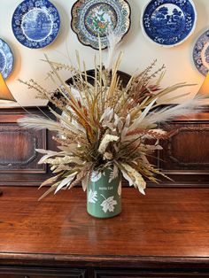 a vase filled with lots of dried flowers on top of a wooden table next to plates