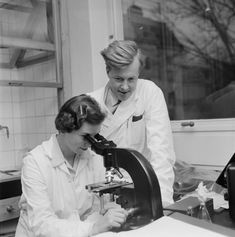 black and white photograph of two women looking through a microscope at something in front of them