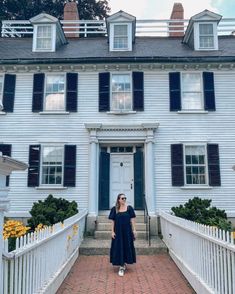 a woman standing in front of a white house with black shutters on the windows