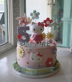 a hello kitty birthday cake with flowers and butterflies on it's top, sitting in front of a window