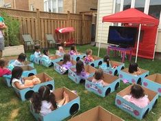 several children are sitting in cardboard cars on the grass with an adult watching tv behind them