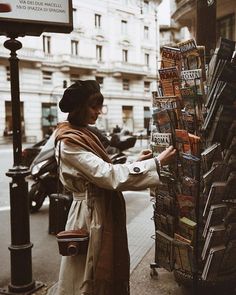 a woman standing next to a pile of books on the side of a street near a building