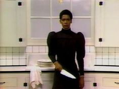 a woman in a black dress standing next to a counter with plates and knives on it