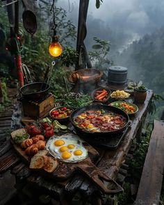 an outdoor table with food on it in the middle of some trees and foggy mountains