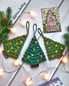 crocheted christmas tree ornaments are displayed on a white wooden surface with lights around them