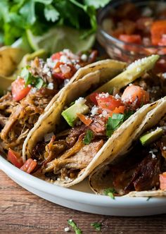 three shredded pork tacos on a plate with salsa and avocado in the background