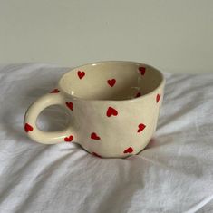 a white cup with red hearts on it sitting on a bed next to a pillow