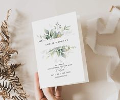 someone holding up a wedding card with greenery on it