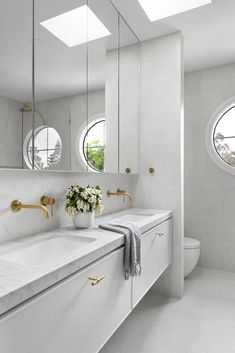 a white bathroom with two sinks and three round mirrors on the wall above them are gold faucets