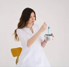 a woman holding a cup and spoon in her hand while standing next to a yellow chair