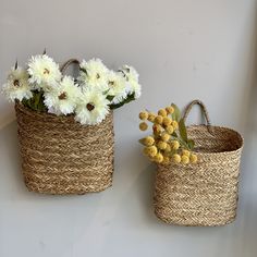 two woven baskets with flowers in them on a white wall next to another basket filled with yellow and white flowers