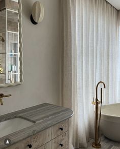 a bathroom with a tub, sink and mirror next to a window that has sheer curtains