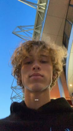 a young man with curly hair standing in front of a building holding a cell phone