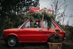 a small red car with presents on the roof and in the passenger seat, sitting next to a basket full of christmas tree branches