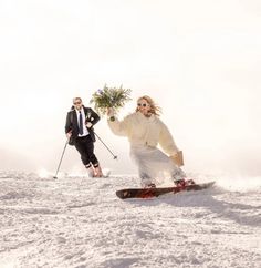 two people on skis in the snow with one holding a small tree and another standing behind them