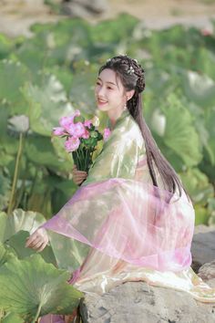 a woman sitting on top of a rock with flowers in her hand next to water lilies