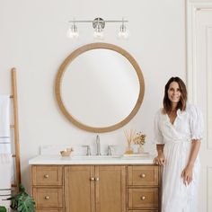 a woman standing in front of a bathroom vanity with a round mirror above it and she is wearing a white robe