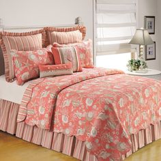 a bed covered in pink and orange comforter next to a white table with lamp on it