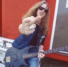 a man with long hair and sunglasses holding a guitar in front of a red wall