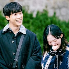 a man and woman standing next to each other in front of green bushes smiling at the camera